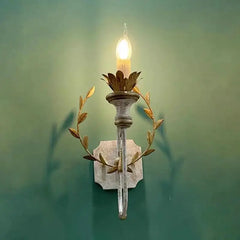 Distressed gold metal wall sconce with a leaf wreath design illuminated against a green wall, casting subtle shadows.