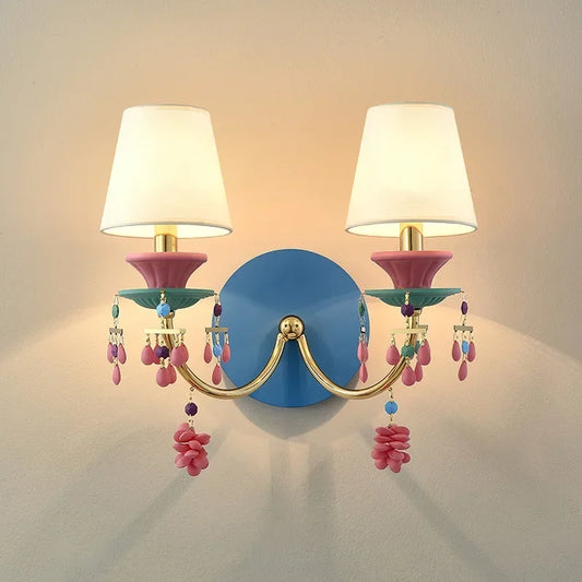 Gold wall sconce with white fabric shades and colorful beaded and flower-like accents, mounted on a blue backing plate.