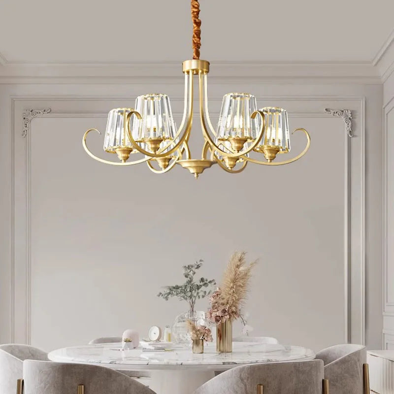A polished gold chandelier with crystal elements is showcased above a dining table with upholstered chairs.