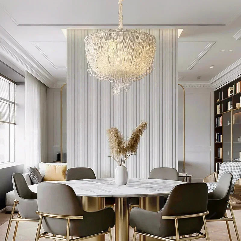 A spacious dining room featuring a polished chrome crystal waterfall pendant light suspended above a round marble table with grey chairs.