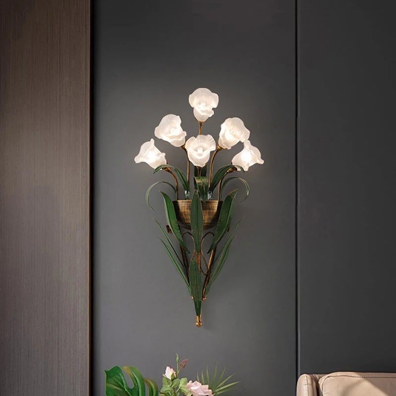 Gold floral wall sconce with clear glass flower shades and green leaf accents mounted on a dark grey wall.