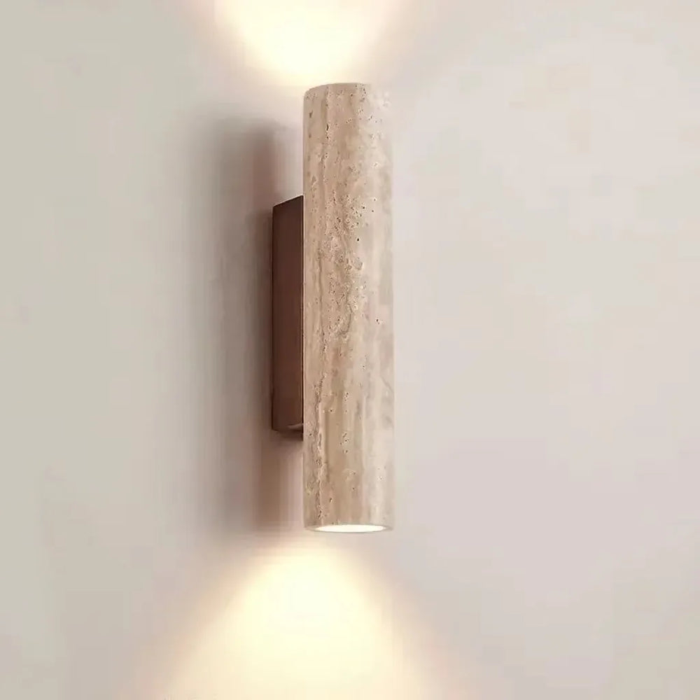 A single cylindrical wall sconce made of textured, light-colored stone, mounted on a dark bracket, emitting light upwards and downwards against a plain wall.