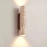 A single cylindrical wall sconce made of textured, light-colored stone, mounted on a dark bracket, emitting light upwards and downwards against a plain wall.
