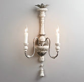 A distressed white wood carved wall sconce with two candle-style lights mounted on a gray wall.