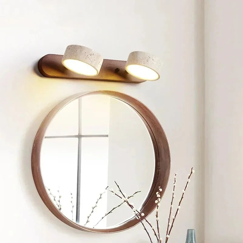 A wall-mounted light fixture with a wooden base and two cylindrical stone shades angled downwards, illuminating a round mirror below.