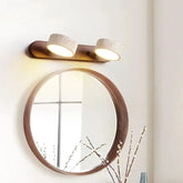 A wall-mounted light fixture with a wooden base and two cylindrical stone shades angled downwards, illuminating a round mirror below.
