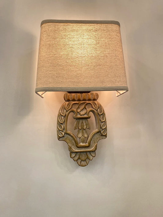 Close-up of a vintage-style wall sconce with a carved wooden base and a light beige fabric shade mounted on a white wall.