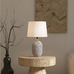A ceramic table lamp with a geometric pattern base and a cream fabric shade, situated on a rustic wooden stump table with a vase of branches.