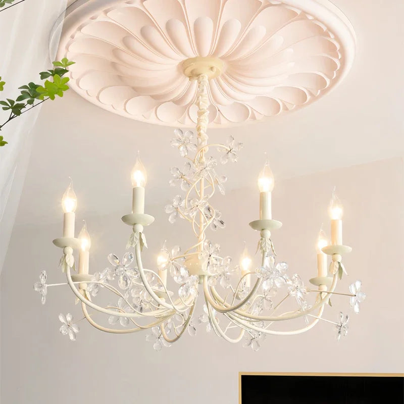 View of a white chandelier with clear crystal flower accents and candle-shaped lights against a white ceiling.