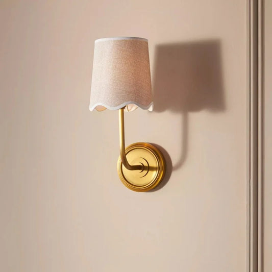 A close-up of a wall sconce with a copper base and a textured fabric shade with a scalloped edge, casting a warm shadow on a neutral wall.
