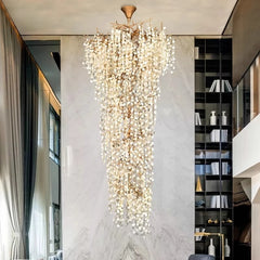 A tall, multi-tiered crystal chandelier with a gold frame illuminates a spacious living area with marble walls and a tall bookshelf.