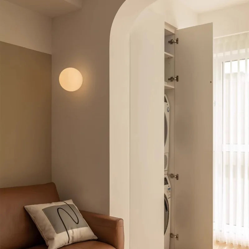 Living room with a brown sofa, round wall light, and open door revealing a laundry area.