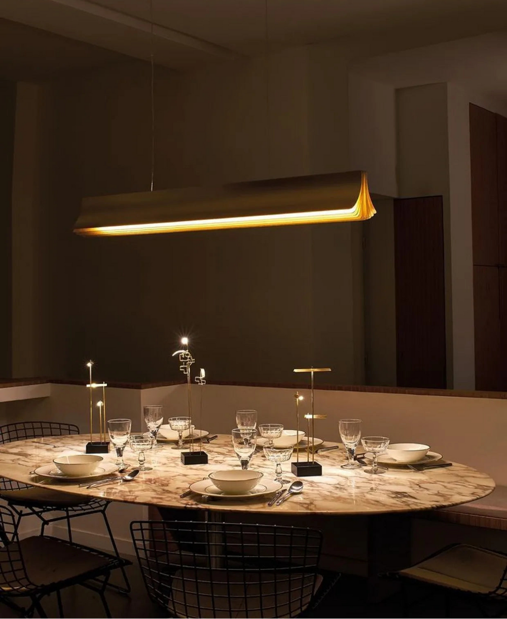 Dining table set with plates, glasses, and cutlery under a modern pendant light in a dimly lit room.