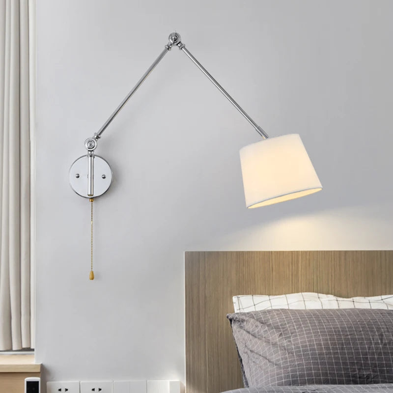 Chrome swing arm wall sconce with a white fabric drum shade extended over a bed.