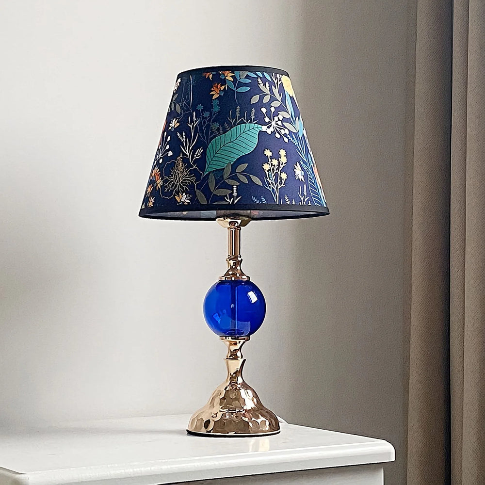 A polished brass table lamp with a conical blue floral fabric shade and a round blue glass orb accent, shown on a white surface.