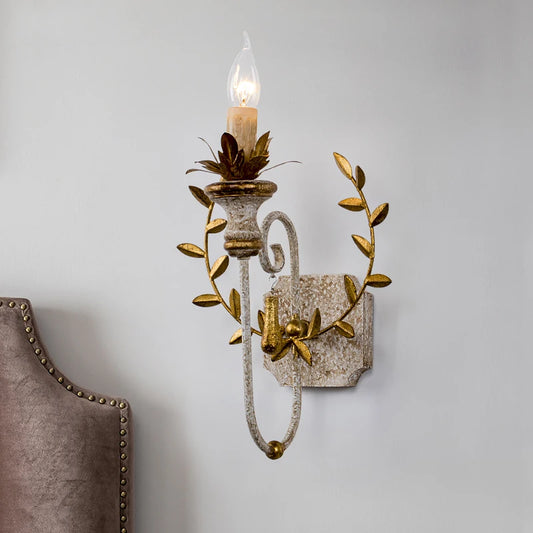 Distressed gold wall sconce with a floral leaf design mounted on a white wall next to a upholstered headboard.