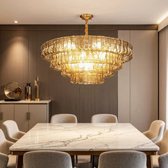 A multi-tiered gold chandelier with amber glass accents hanging over a rectangular marble dining table with grey fabric chairs.