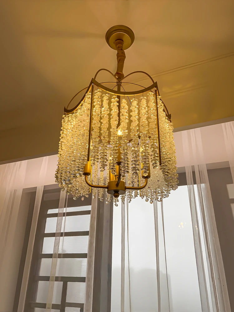 A gold crystal chandelier with cascading droplets hanging in front of a window with sheer white curtains.