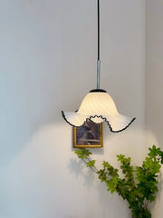 Pendant light with white lampshade and black trim hanging against a plain wall with a plant and framed picture.