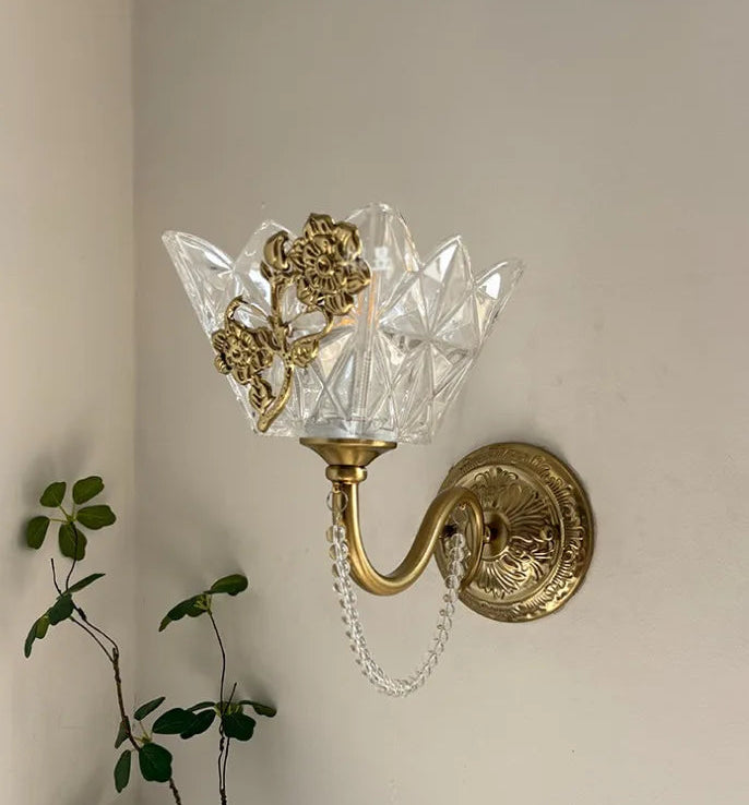 A gold wall sconce with a clear, faceted glass shade adorned with a floral metal accent, mounted on a neutral wall with a plant in the foreground.