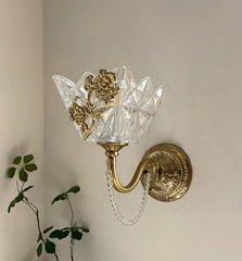 A gold wall sconce with a clear, faceted glass shade adorned with a floral metal accent, mounted on a neutral wall with a plant in the foreground.