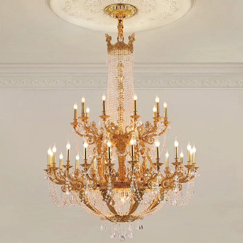 Close-up of a gold tiered chandelier with crystal accents and candelabra-style arms, illuminated against a white ceiling and wall.