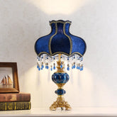 A decorative table lamp with a scalloped blue fabric shade featuring gold trim and hanging crystal and blue beads. The base is gold with ornate detailing and a bulbous blue ceramic section.