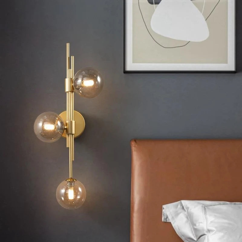 A brass wall sconce with clear glass globes displaying lit bulbs, positioned near a leather headboard and bedding in a bedroom setting.