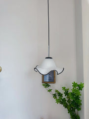 Pendant light fixture with white lampshade and black accents hanging against a plain wall with a plant in the foreground.