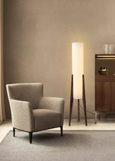 A minimalist floor lamp with a black walnut base and a tall, cylindrical white fabric shade, standing next to a textured armchair in a room with muted walls.