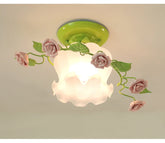 A close-up view of a green base ceiling light adorned with pink ceramic roses and green leaves, emitting a soft glow from its frosted glass shade.