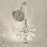 Close-up of a wall-mounted light fixture with a faceted crystal flower design and a polished chrome arm, casting prismatic light patterns on a white wall.