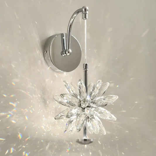 Close-up of a wall-mounted light fixture with a faceted crystal flower design and a polished chrome arm, casting prismatic light patterns on a white wall.