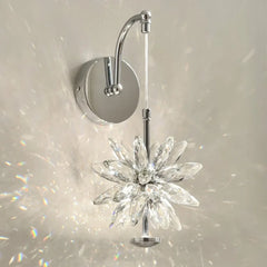Close-up of a wall-mounted light fixture with a faceted crystal flower design and a polished chrome arm, casting prismatic light patterns on a white wall.