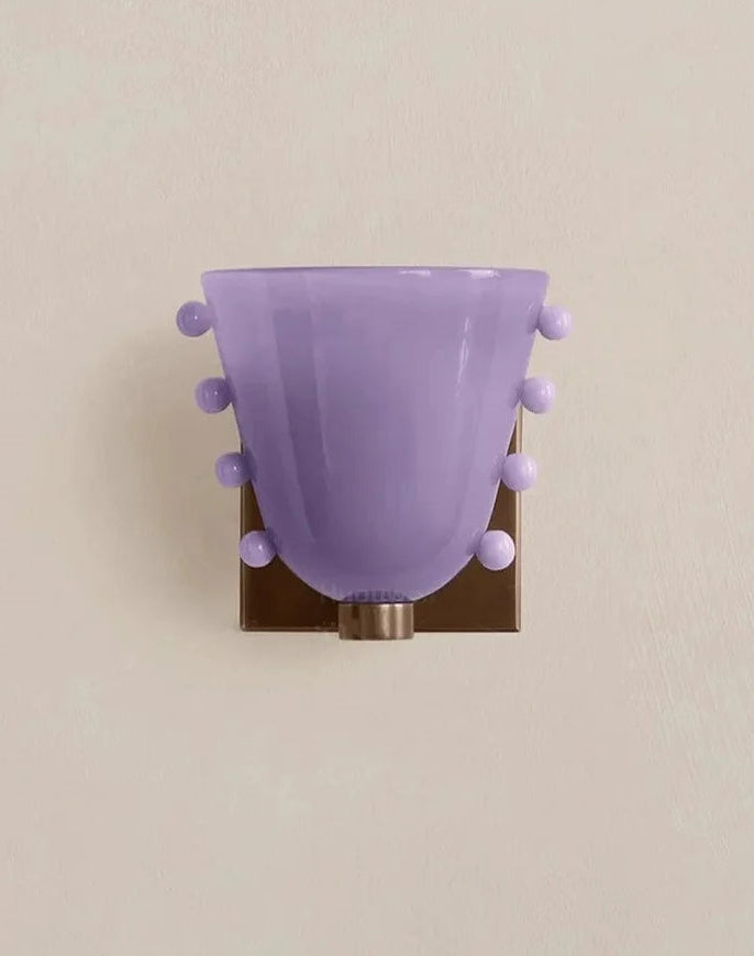 Decorative wall light fixture with a purple glass shade and pom-pom details on a beige background