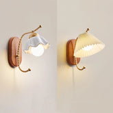 Two wall sconces side-by-side, one with a white ceramic flower shade and the other with a pleated cream fabric shade, both mounted on wooden bases with gold arms.