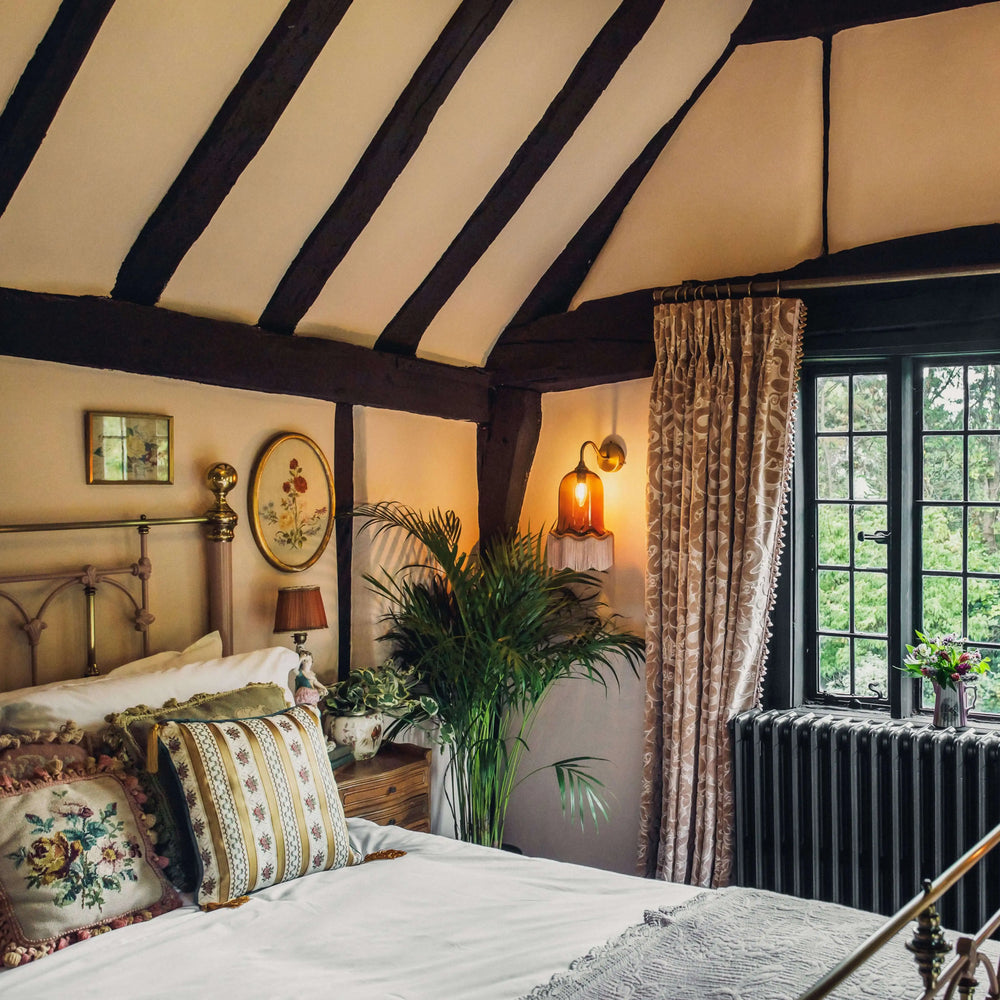 A wall-mounted brass sconce with a bell-shaped glass shade and tassel trim, illuminating a cozy bedroom with a canopy bed and dark wooden beams.