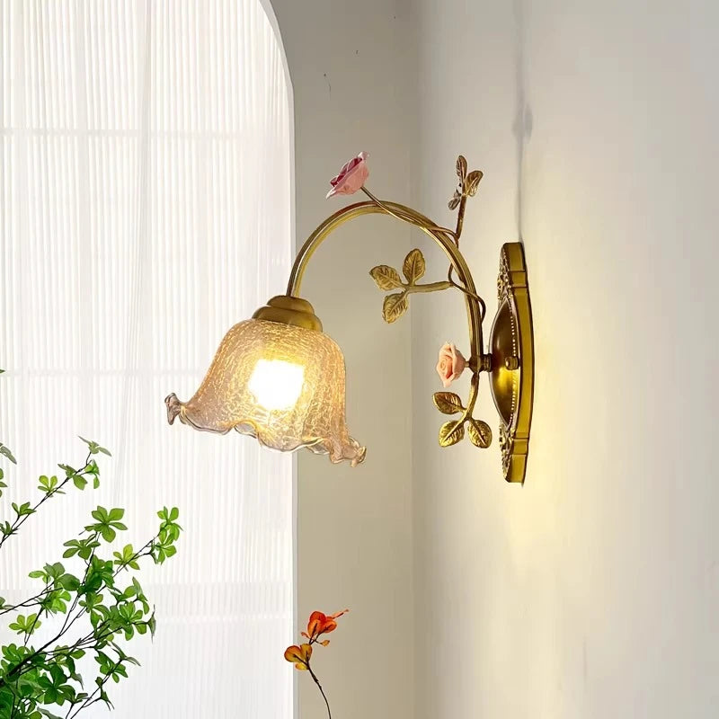 A gold floral wall sconce with a fluted glass shade is illuminated against a white wall, with green foliage visible in the foreground and a sheer curtained window in the background.