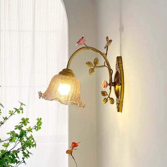A gold floral wall sconce with a fluted glass shade is illuminated against a white wall, with green foliage visible in the foreground and a sheer curtained window in the background.