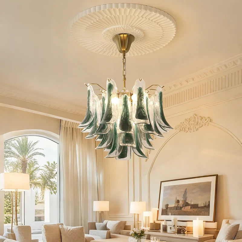 A gold-tone tiered chandelier with cascading glass shades, installed in a well-lit living room with large windows and elegant furnishings.
