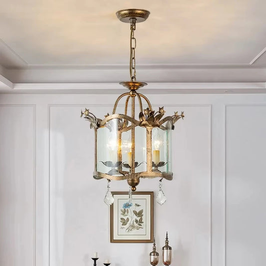 A gold chandelier with a distressed finish, glass cylinder shades, and crystal teardrop pendants hanging from a ceiling.