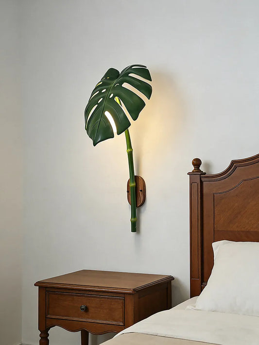 Green monstera leaf wall sconce with a faux bamboo stem mounted on a white wall next to a wooden nightstand and bed.