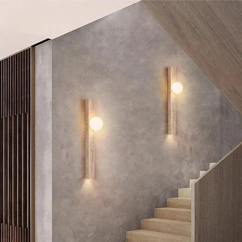 Two travertine stone wall sconces with frosted globes mounted on a textured wall next to a wooden staircase.