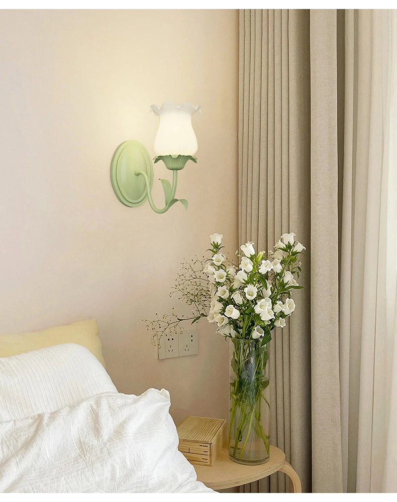 A single green floral wall sconce with a frosted glass shade, installed on a wall next to a bed and a vase of flowers.