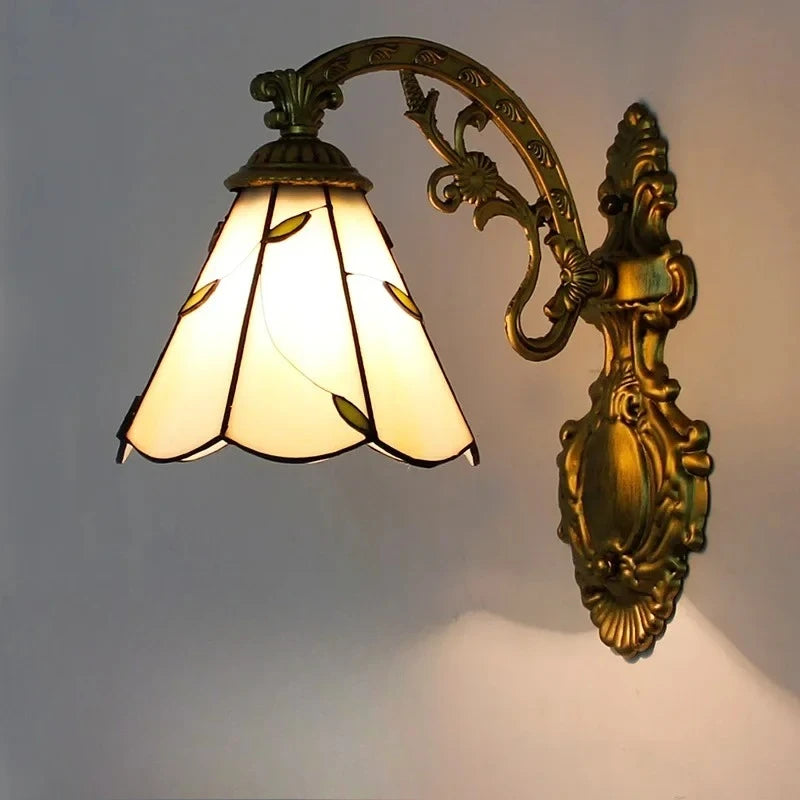 A wall-mounted light fixture with an antique gold metal arm and a tapered white stained glass shade with green leaf accents, lit from within.