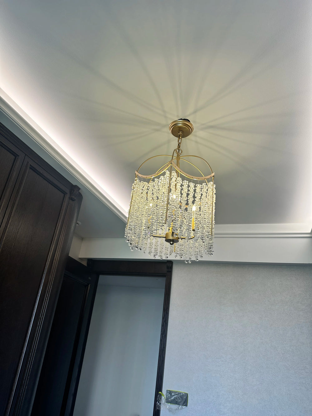 A gold crystal chandelier with cascading droplets casting shadows on a white ceiling and wall in a modern room.