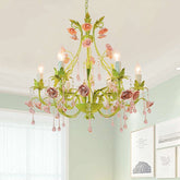 A green chandelier with pink and purple roses, green leaves, and dangling pink crystals, illuminated by candle-style lights.