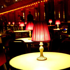 An indoor scene with multiple tables, each featuring a lit burgundy pleated fabric shade table lamp, creating a warm atmosphere.