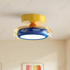 Close-up of a modern flush mount ceiling light with a yellow top, orange accent, and a blue and clear ring diffuser with a white light source inside, mounted on a white ceiling.