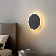 A single black circular wall sconce with a warm halo effect mounted on a grey wall next to a bed.